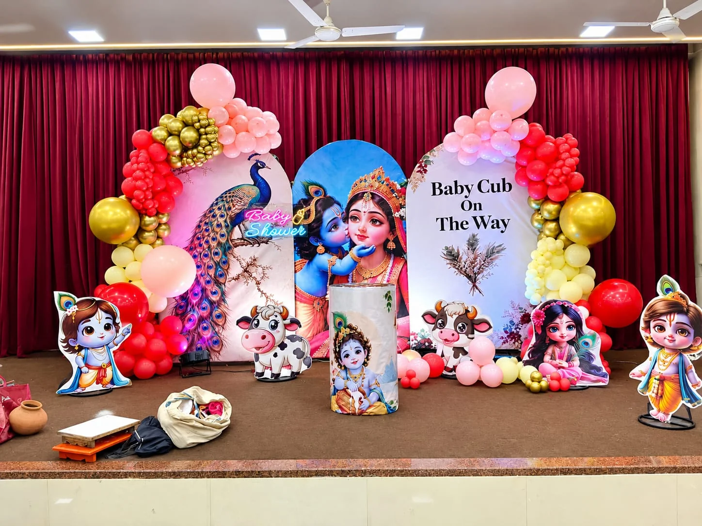 Krishna-themed baby shower stage with three arch panels and crimson, blush and gold balloon garlands