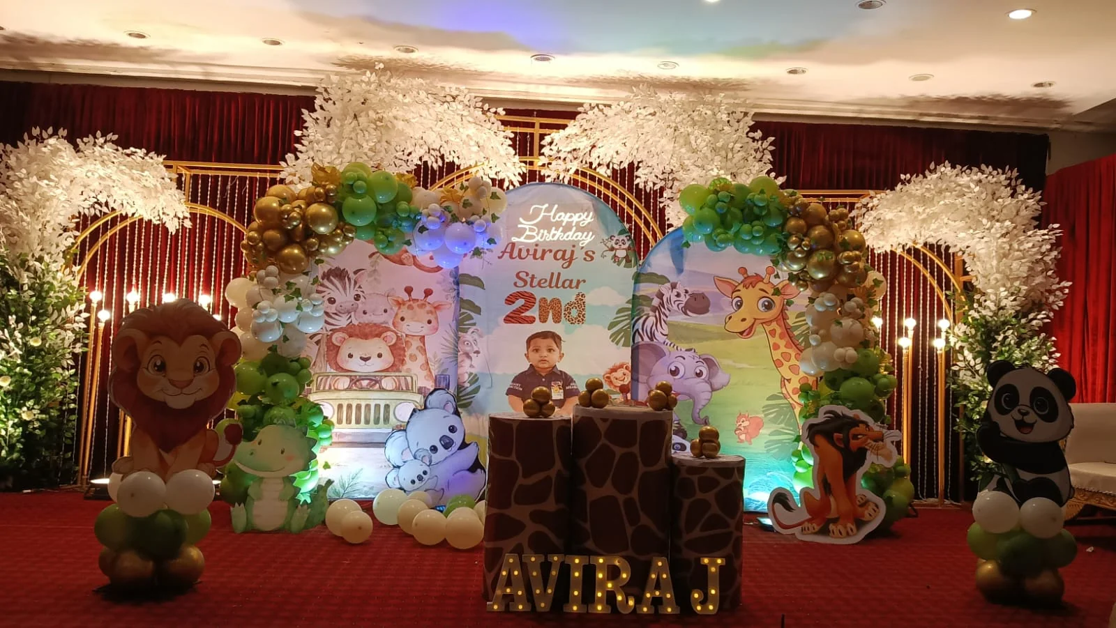 Safari-themed birthday stage with arch panels, green and gold balloon garlands, white foliage trees and animal cut-outs